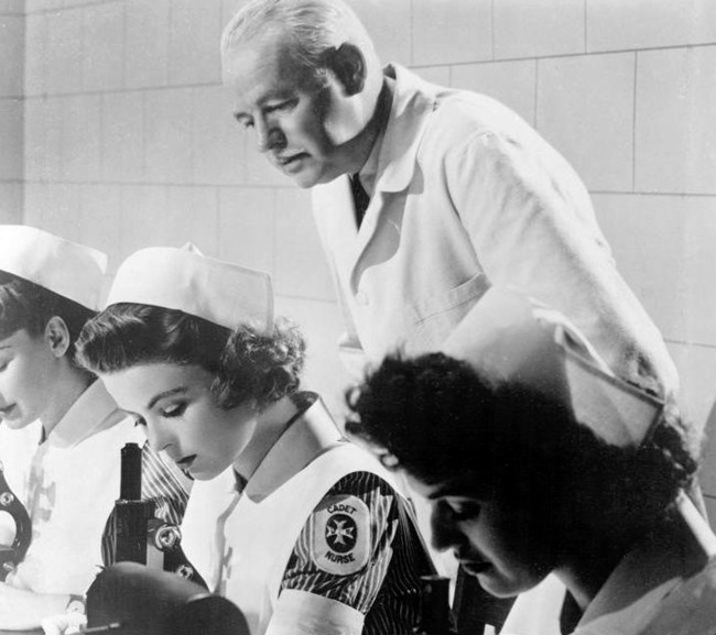 A still from the short film "Reward Unlimited" (1944), which promoted the Cadet Nurse Corps. https://en.wikipedia.org/wiki/Cadet_Nurse_Corps#/media/File:Reward-Unlimited-5.jpg. Black and white of three women in nurse caps with a Cadet Nurse Corp Patch looking into microscopes. An older man in a white coat looks over their shoulder.