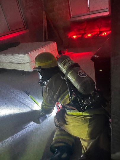 Firefighters use a flashlight and crawl through a dark house.