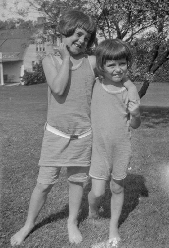 A black and white photo of two small girls in summer rompers and bob haircuts smiling and posing barefoot with their arms around each other on a large grassy lawn in front of a cottage and trees.