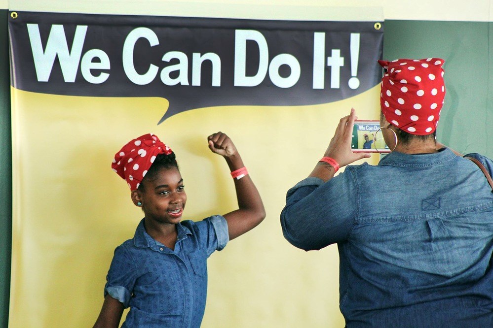 Color photo of a young African American girl in blue coveralls and red bandana with white polk dots having her picture taken by an adult in blue coveralls with matching bandana in front of a speechbubble reading "We Can Do It!"