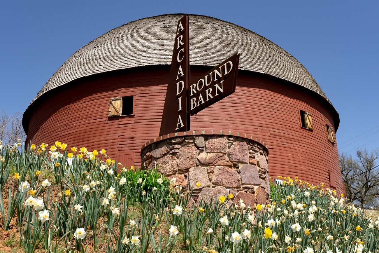 A round building with a rounded roof, surrounded by flowers. A sign reads "Arcadia Round Barn".