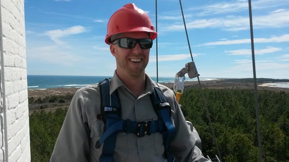 Sabol wears a hardhat, glasses and harness on the Cape Lookout Lighthouse with trees and the sea in the background below him.