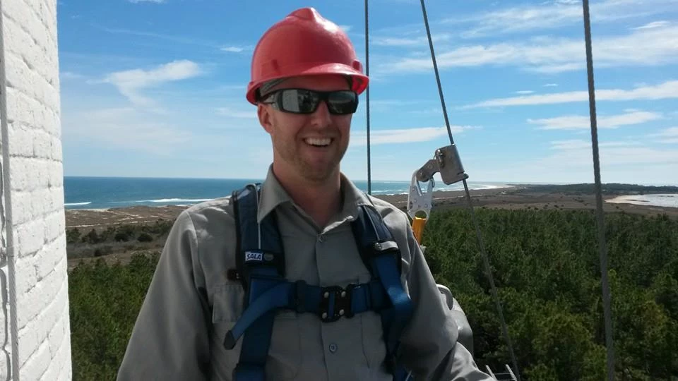 Cape Lookout Lighthouse Exterior Painting Sabol wears a hardhat, glasses and harness on the Cape Lookout Lighthouse with trees and the sea in the background below him.