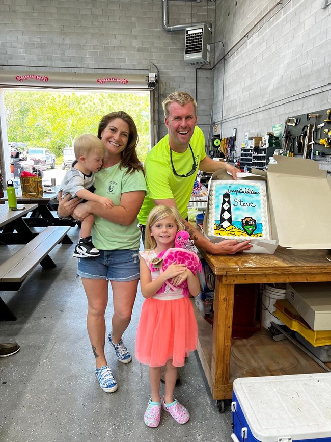 Steve stands inside a building next to family holding up a farewell cake.