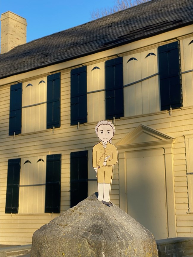 A cartoon illustration cutout of Alexander Hamilton is placed in front of a yellow house with many windows, with black shutters.