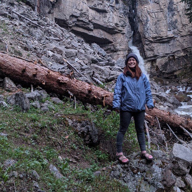Woman stands on rocky slope  with a waterfall streaming from a cliff behind her.