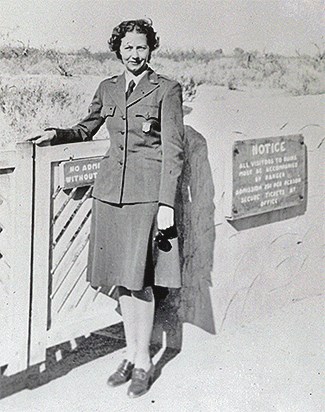 Sallie Pierce Brewer Negative A woman wearing an NPS uniform with her badge pinned to her left side chest pocket is seen with her right arm on a gate. She faces left and holds sunglasses in her left hand.