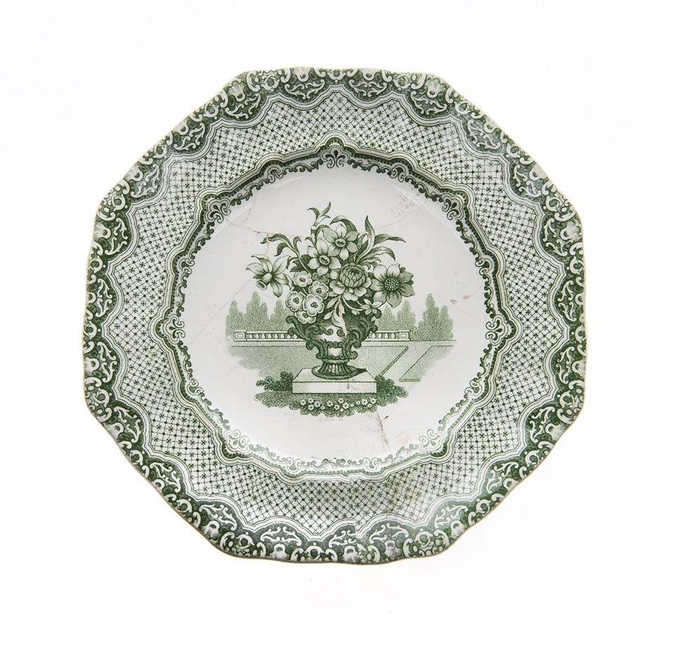 An octagonal plate, white with a green transfer print design. The design shows a vase full of flowers surrounded by a border of stars and scrolls.