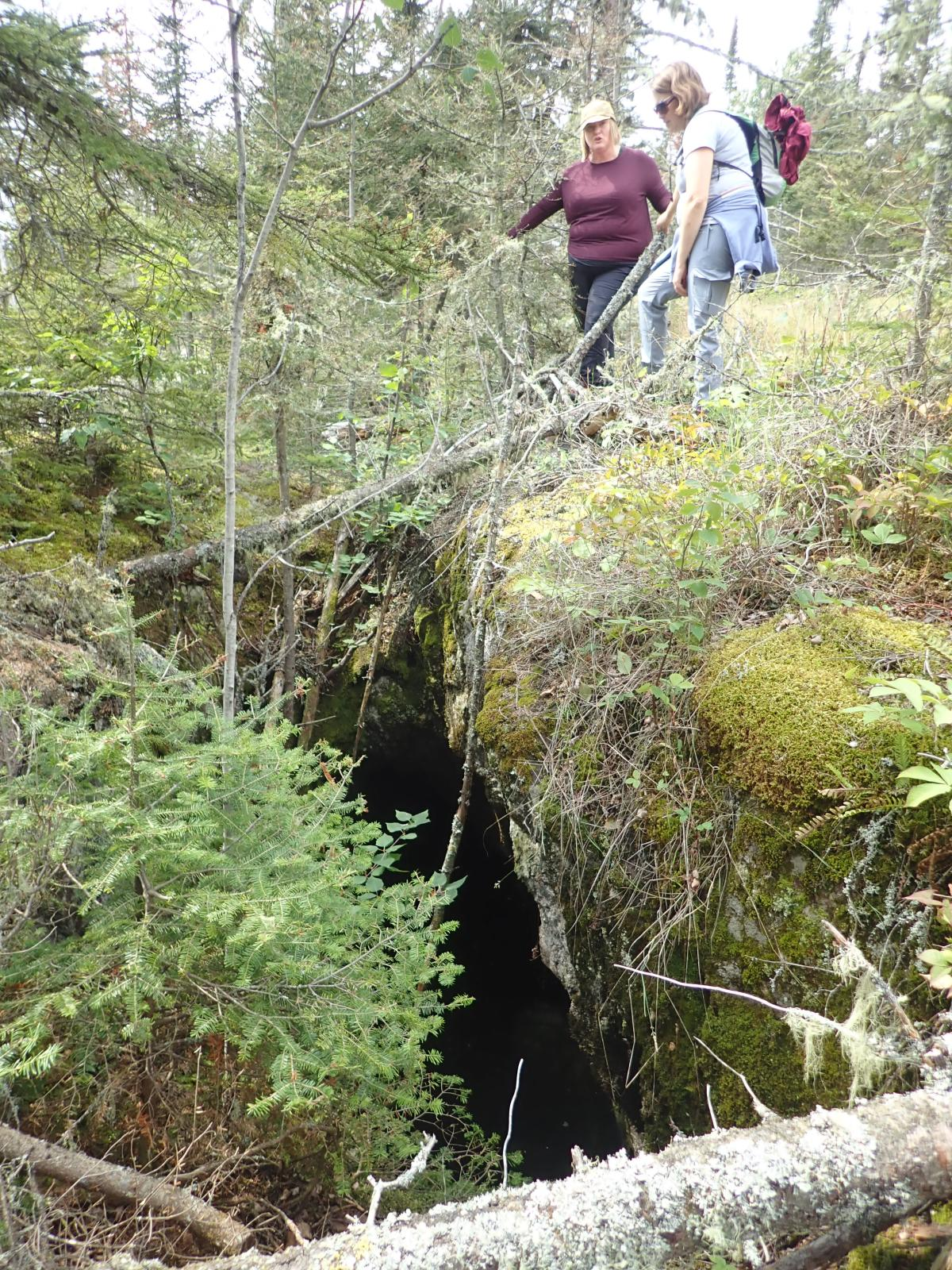 Two people stand over an elongated hole in the moss-covered hard rock with coniferous green trees surrounding.