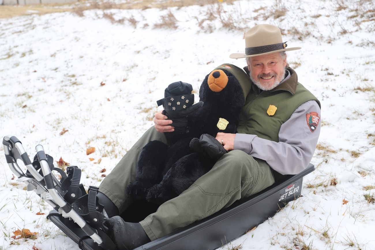 Craig Olsen sitting on a sled with snowshoes, holding a stuffed bear keeping their paw warm with gloves.