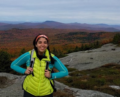 A woman in athletic wear and a puffy florescent jacket stands in front of a fall foliage landscape