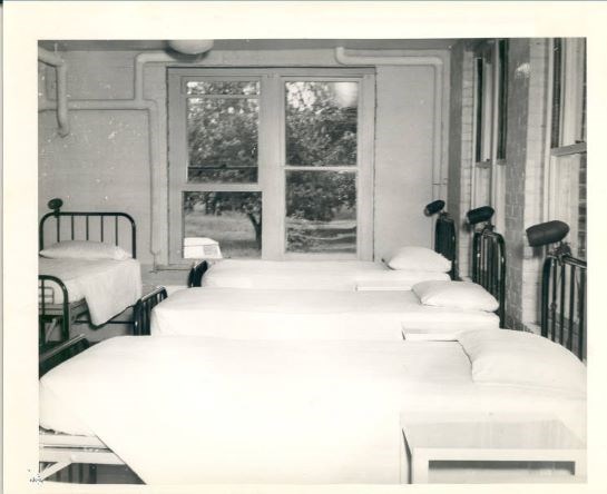 Black and white image of a row of made beds. A large window in the back shows a tree outside. Whitewashed walls show exposed piping.
