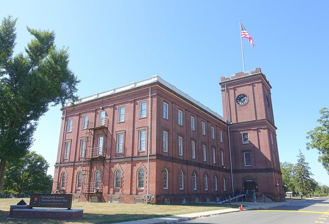 Red brick three story building. A red brick tower is attached on the right with a clock face and an American flag flying on the top. National Park Service sign visible in the foreground.