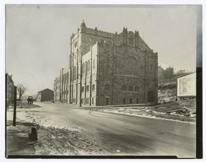 Black and white photo of large brick building with gothic features on a wide, muddy road.