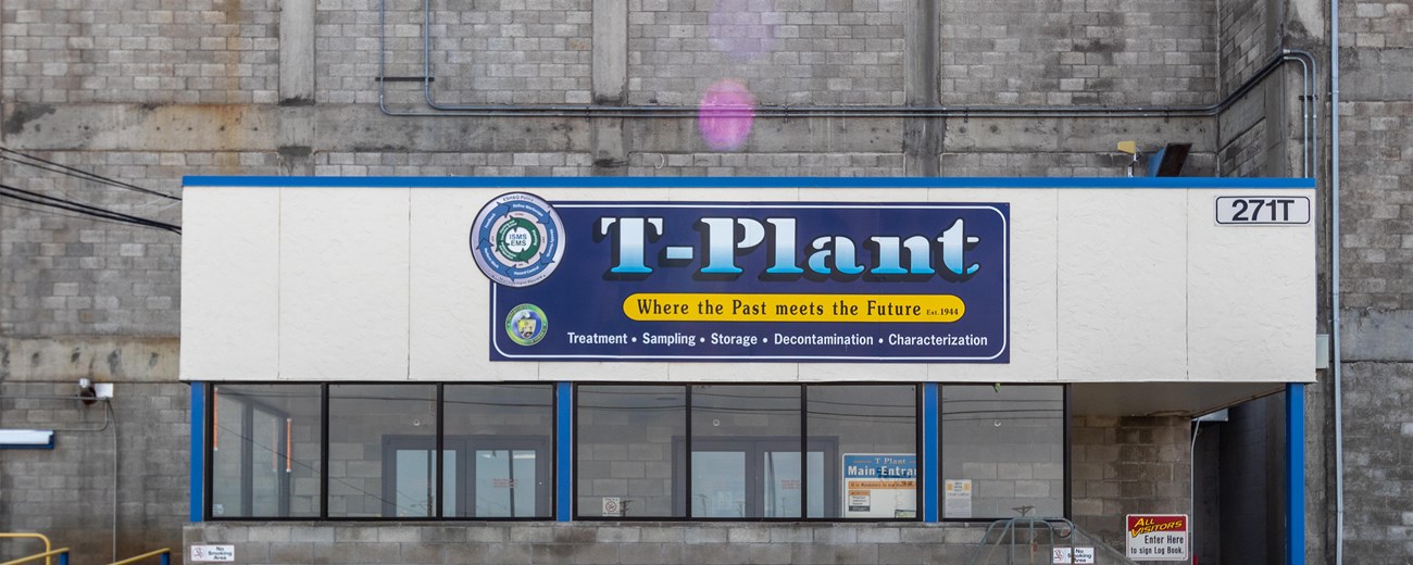 An image of a large sign on a concrete building reads “T Plant” with a blue background. Glass panes are directly below the sign.
