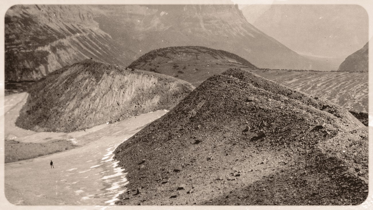 Black and white photograph of an alpine pass with a small glacier and a moraine in the foreground. A person is on the left in the distance.