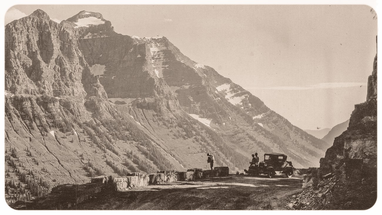 Black and white image of a mountain road with a parked car and people.