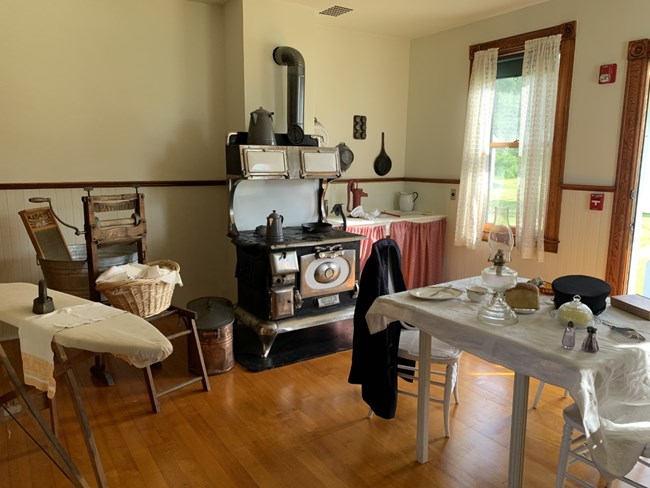 An example of a kitchen in the 1920's with an ironing board, laundry basket, black metal wood fired stove, and a table set with dishes and a lantern.