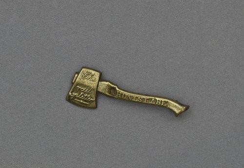 A small gold pin is shaped like an axe. Raised text is printed on the blade reading “The rail splitter,” and handle reading “Honest Abe.”