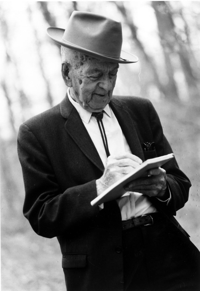 Freeman Tilden in suit and hat stands outside writing in a notebook