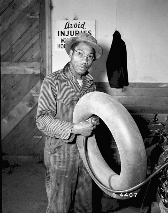Black and white photo of an African American man in coveralls and a hat holding a tire