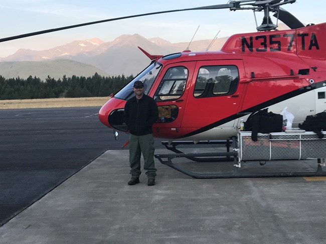Todd stands in front of a helicopter while on a firefighting assignment in Glacier National Park. Smiling man stands in front of red helicopter with mountains in the background.