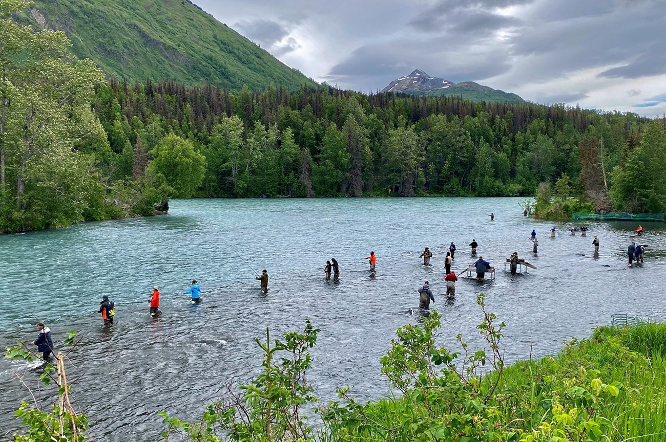 Chugach National Forest A verdant forest surrounds a scenic river filled with people fishing. Snow-capped mountains are in the background.