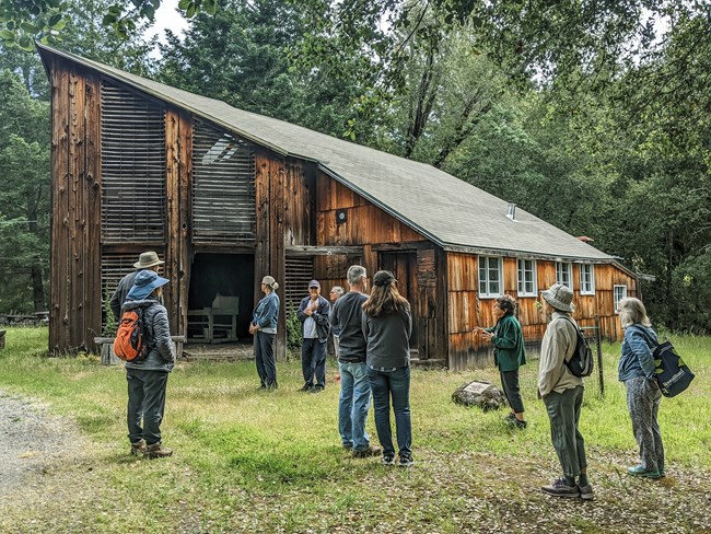 Group of people stand in circle in front of barn in woods