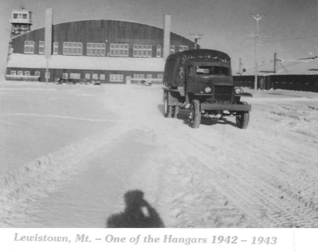 Black and white photo of a large military truck driving in front of an airport hangar