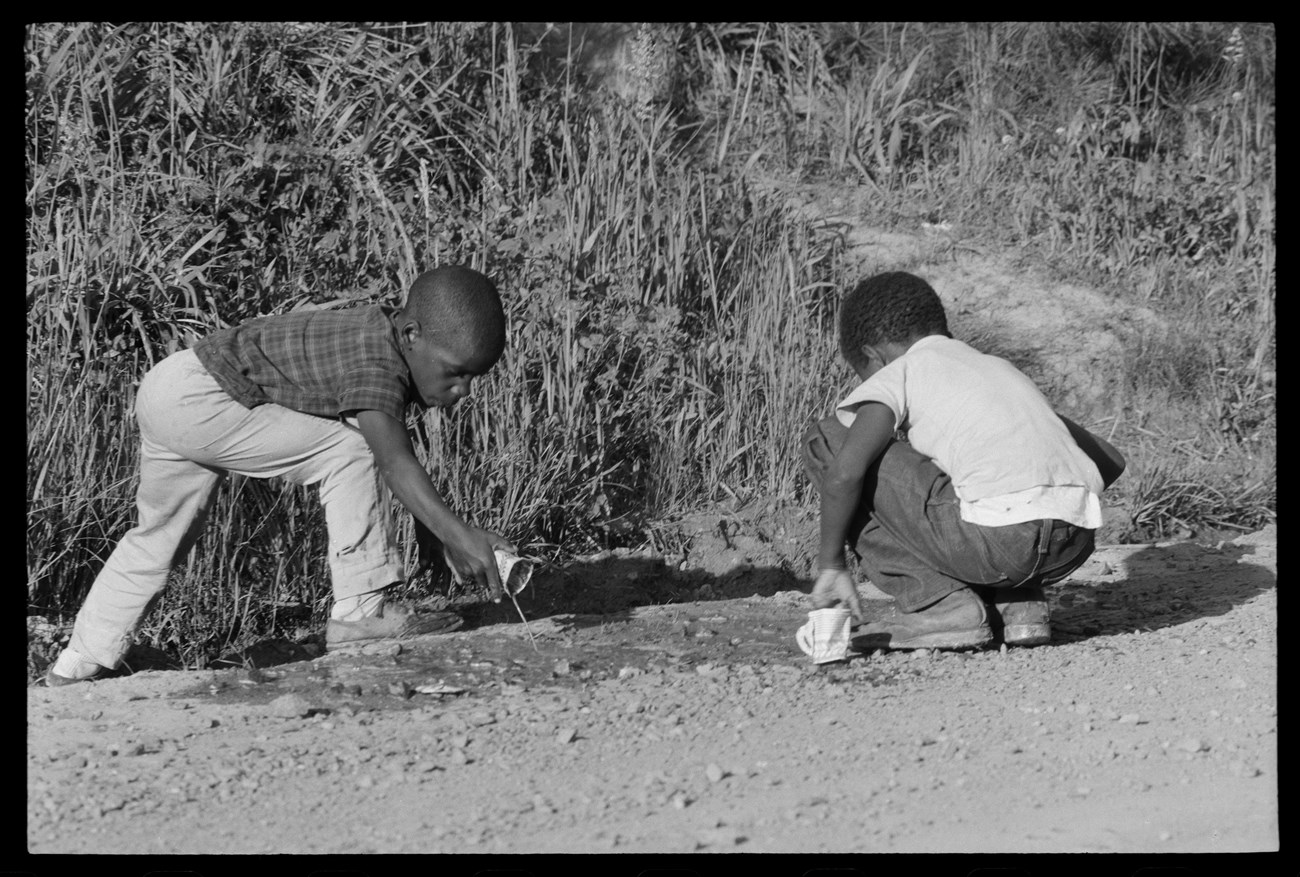 Two African American boys play in a ditch between a field and a road.