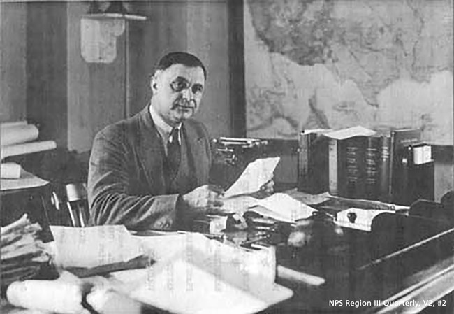 Arno Cammerer sitting at desk with paper in hand looking at camera