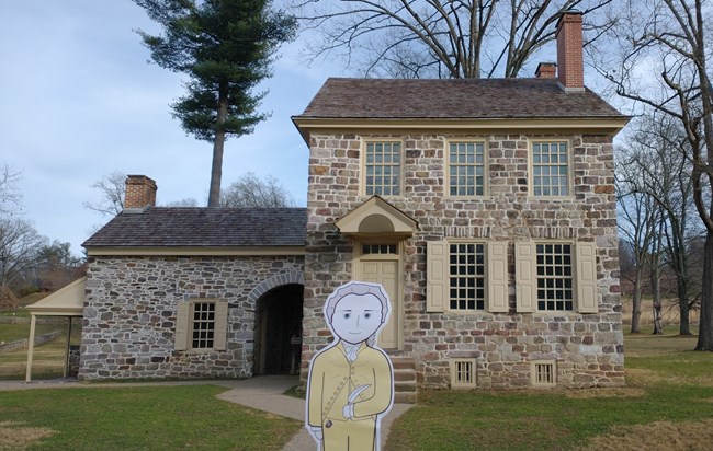 A cartoon illustration cutout of Flat Hamilton in front of a stone house.