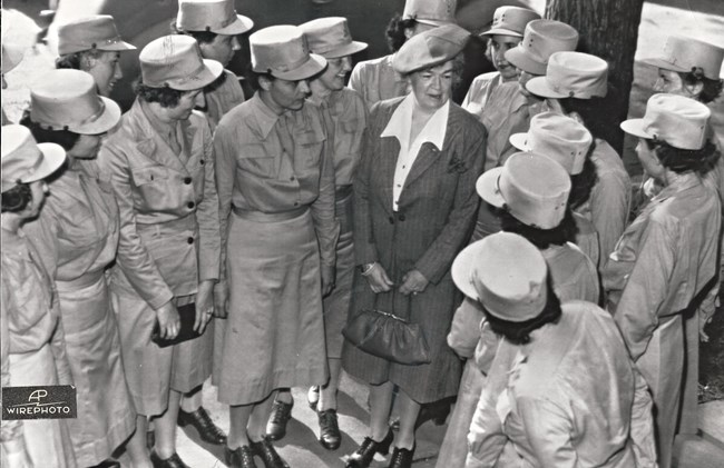 WAACs meet Rep. Edith Nourse Rogers Women wearing cylindrical military caps and collared dresses with skirts below the knee stand side by side in a U shape to meet an older woman. The woman wears a dark, pin-striped dress with a white collared shirt and holds a small purpose.