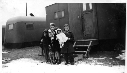 woman crouched down smiling with arms around two children in front of trailer housing