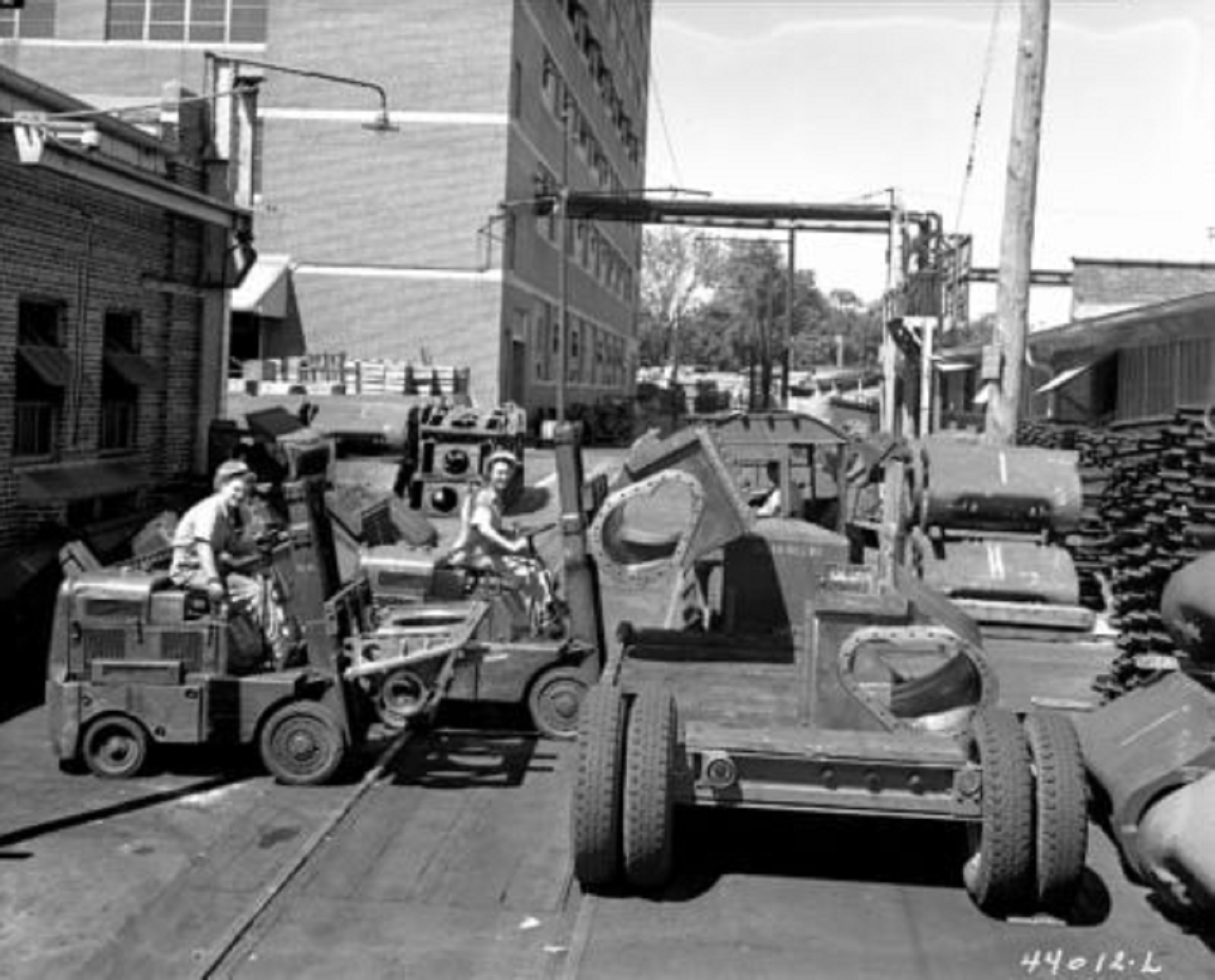 black and white photo of two women driving equipment outdoors at a large manufacturing site.