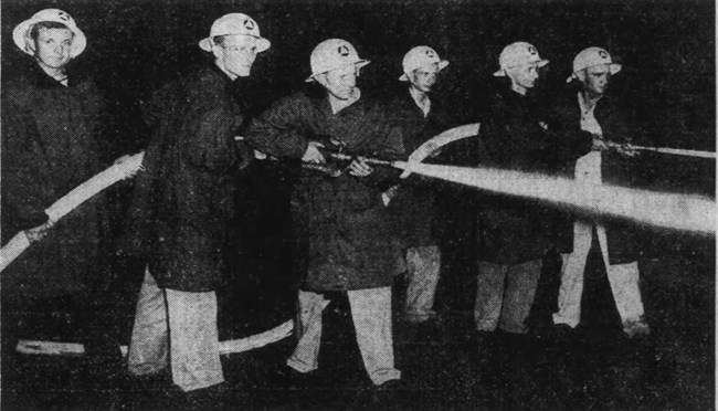 Two groups of three men direct fire hoses that are blasting water at a high pressure. They are wearing civil defense helmets and heavy fireman’s coats.