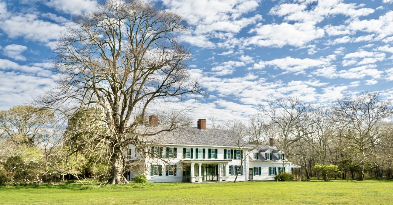 A clapboard white house with more than a dozen front windows and a columned portico surrounded by forest and a bright green lawn.