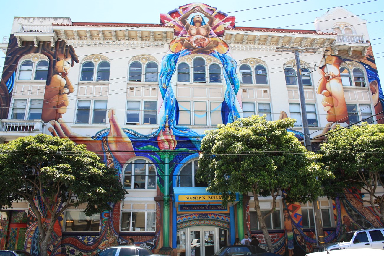 The Women's Building, 18th Street facade White multi-story building with arched windows covered in mural depicting central figure of seated pregnant woman with butterfly wings over falling water flanked by two faces in profile facing one another