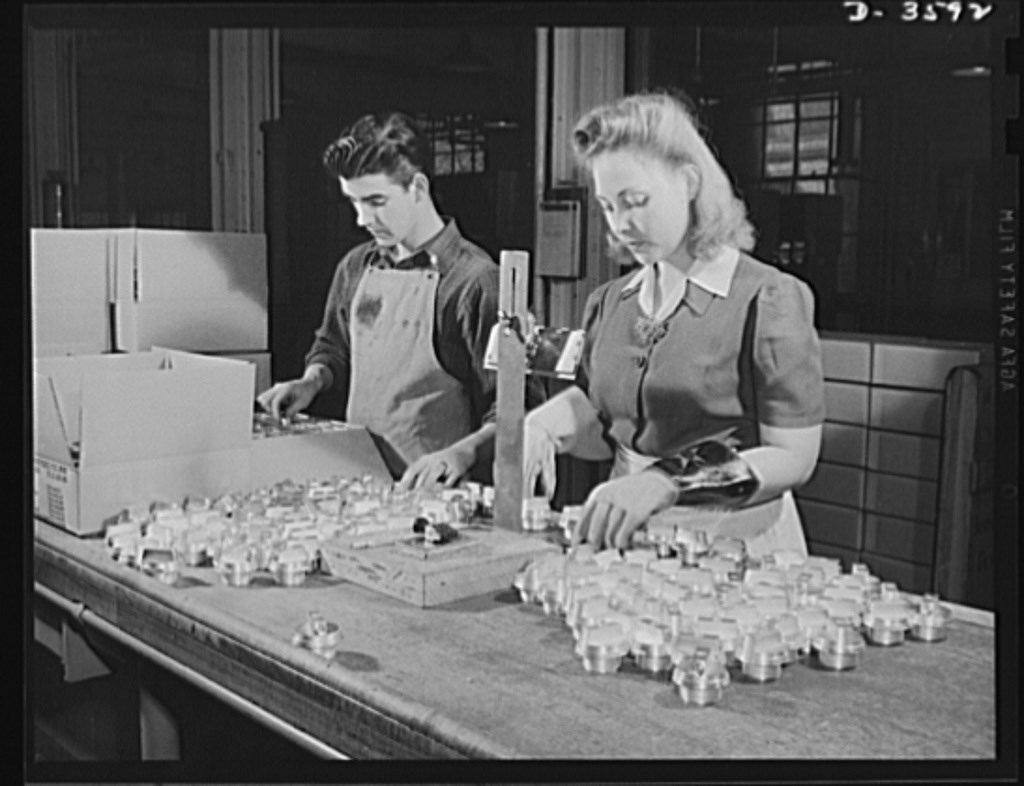 Dark-haired white man and blonde woman test and inspect parts for ammunitions shells. The man wears an apron and the woman wears thick work gloves.