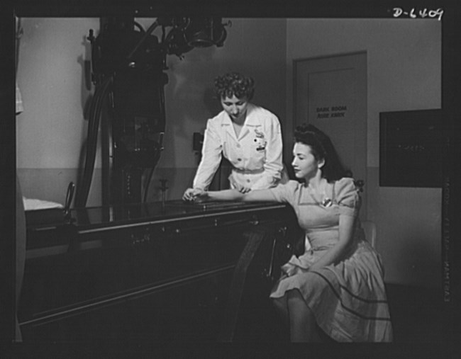 X-ray at North American Aviation hospital. Photo by Alfred T. Palmer, October 1942. Courtesy of the Library of Congress Prints and Photographs Division, LC-USE6-D-006409 (b&w film neg.) Black and white photo of a uniformed female nurse positioning a woman’s right wrist on an X-ray machine. The woman is seated next to the machine with her right arm propped up onto it.