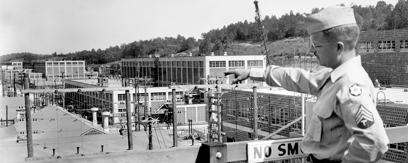 A man in military uniform stands on a balcony pointing with his right arm towards a large industrial complex directly below him.