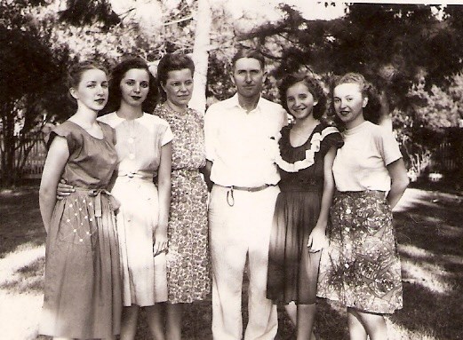 Five women in dresses stand with one man in shirt in pants, outside.