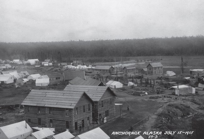 a historic scene of wooden buildings and white tents clustered in front of a tree-lined forest.