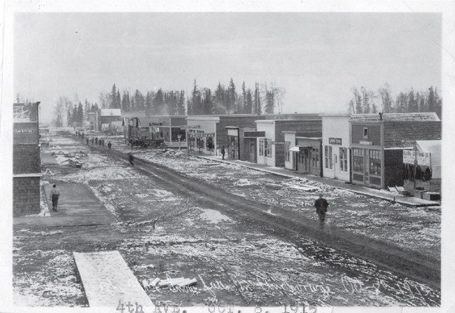 a historic scene of simple, boxy buildings lining a wide dirt street.