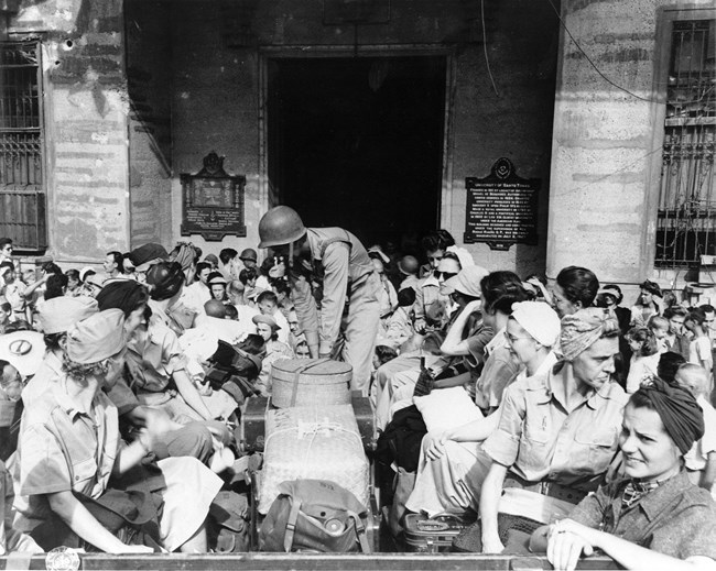 Black and white photo of a crowd of women around a truck with a man in an army helmet in the middle loading boxes.