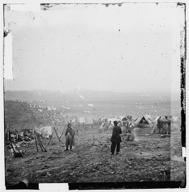 A black and white photograph depicts two men standing on a hilltop cleared of all trees and foliage. To the left are guns arranged in upright tripods. In the background are peaks of white tents where Union soldiers camped