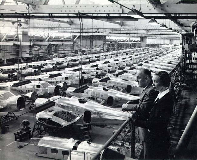 Warehouse with rows of planes. A white man and woman look down over railing at right.