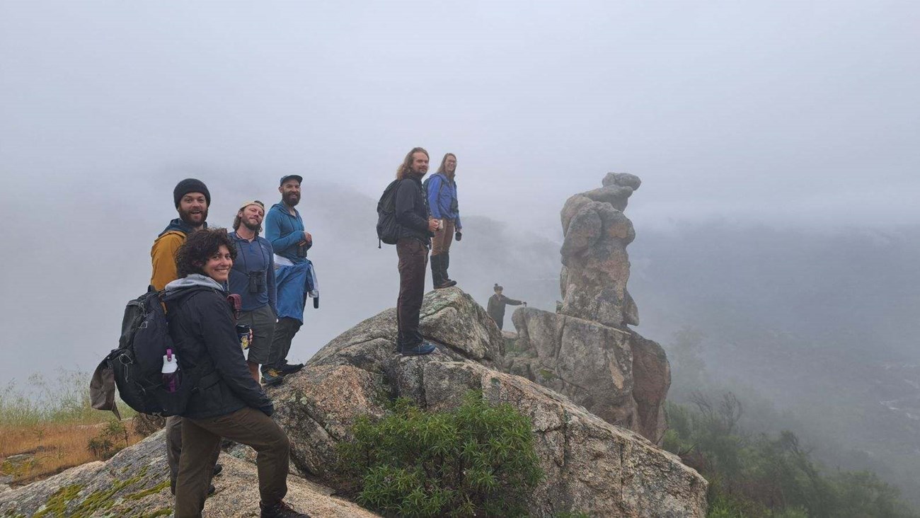 Seven people stand along the top of a granite rock outcrop on a foggy day.