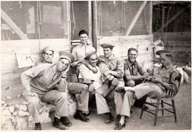 Group of six men, seated, in uniform, outside wooden building.