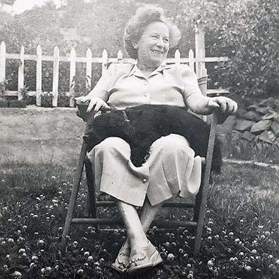 Frieda Fromm-Reichmann sits in a lawn chair with her dog – a small black cocker spaniel - in her lap. She is a middle-aged white woman in a plain, light-colored dress. She is looking to the left and smiling broadly.